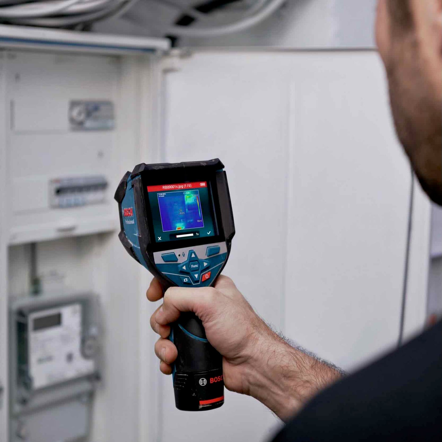 Ein Elektriker führt eine detaillierte Prüfung des Sicherungskastens mit der Bosch GTC 600 C Professional Wärmebildkamera durch. Auf dem Display sieht man einen roten Kreis, der auf eine überlastete elektrische Sicherung hindeutet, während der Rest des Sicherungsschranks in Ordnung und blau dargestellt.