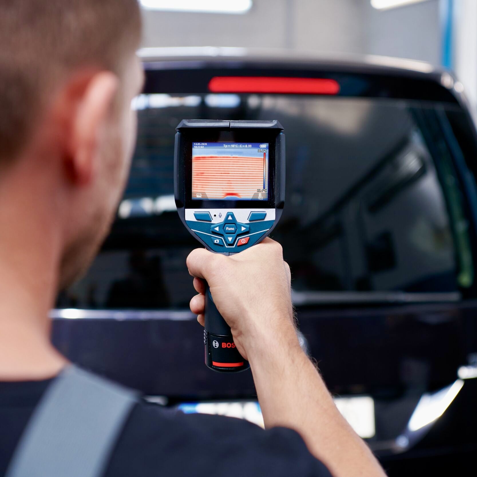 Ein KFZ-Mechatroniker hält die Bosch GTC 600 C Professional Wärmebildkamera und prüft die beheizbare Heckscheibe eines Fahrzeugs. Das Display zeigt ein detailliertes Wärmebild der Heizdrähte zur Funktionskontrolle. Professionelle Thermografiekameras mieten bei: LeihTronik.de